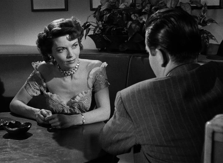 Dorothy Green, as Lucy Chapman, sits across from Glenn Ford’s character in a dimly lit restaurant. Wearing a lace-trimmed dress and a bold necklace, she leans slightly forward with a defiant expression — a crucial moment of revelation in the film’s early investigation.