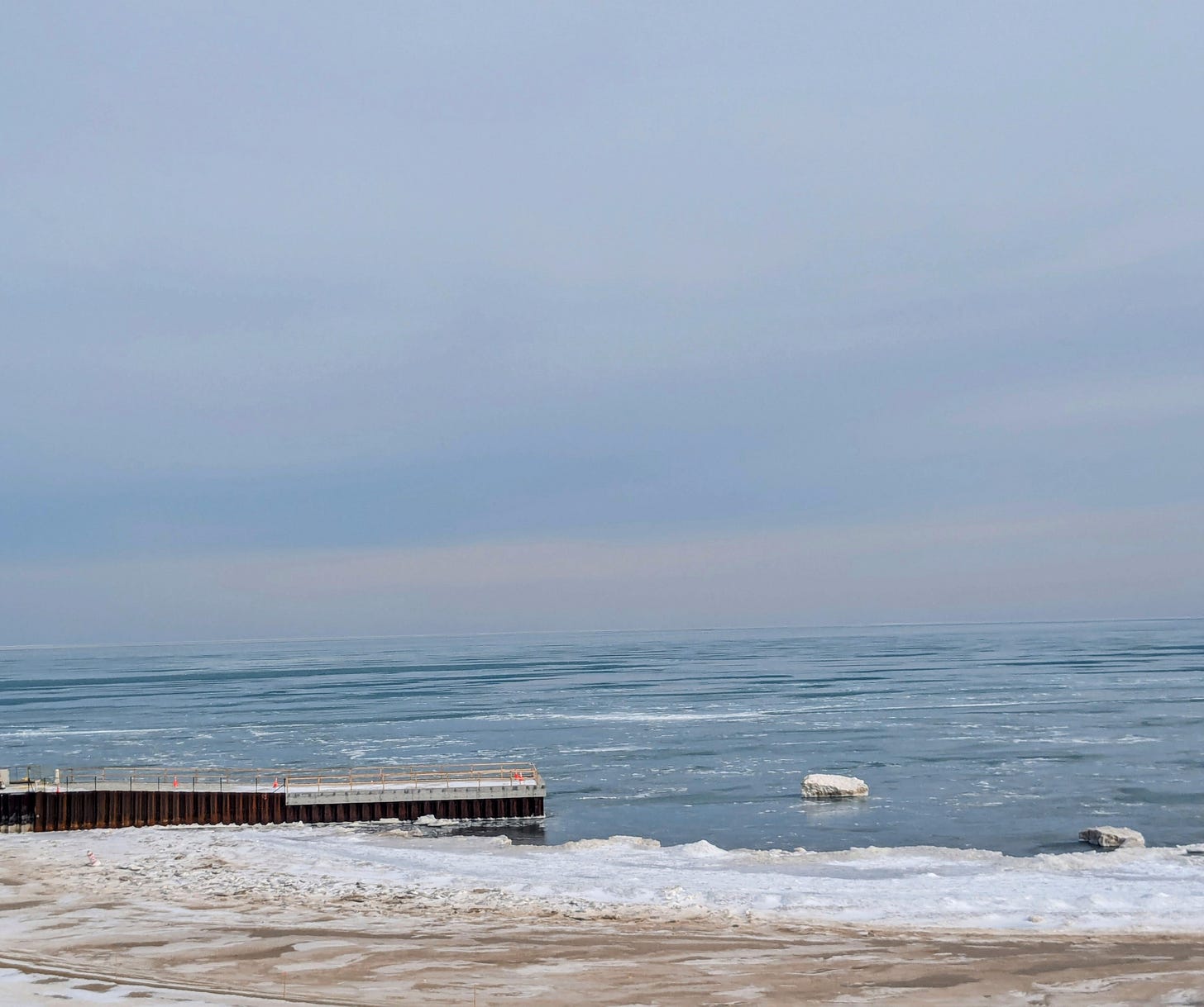 A photograph of Lake Michigan from a beach covered in sand and snow. Ice floats on the water and a wooden pier extends out from the shore.