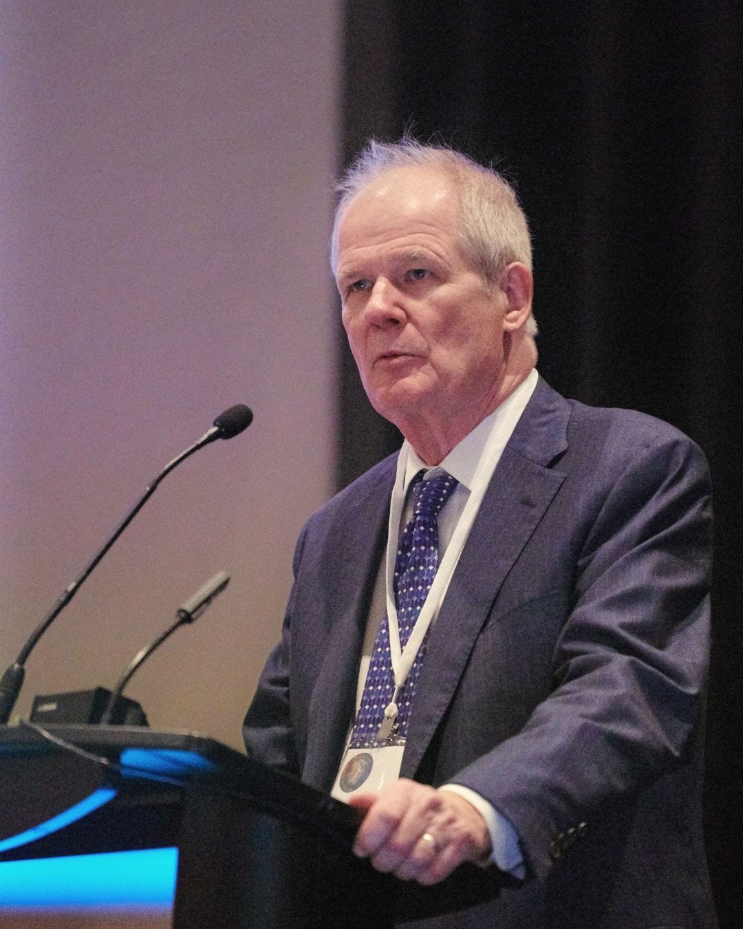 An elderly man in a suit speaks at a podium with a microphone, conveying confidence. He wears a conference badge against a dark background. An elderly man in a suit speaks at a podium with a microphone, conveying confidence. He wears a conference badge against a dark background.