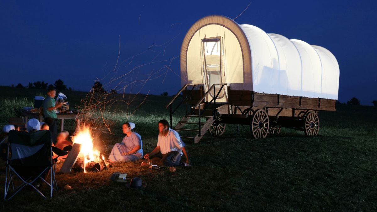 People sit around a campfire next to a covered wagon at dusk, some opting out of the warmth to tend cooking supplies on the ground, while the softly illuminated wagon glows in the evening light. People sit around a campfire next to a covered wagon at dusk, some opting out of the warmth to tend cooking supplies on the ground, while the softly illuminated wagon glows in the evening light.