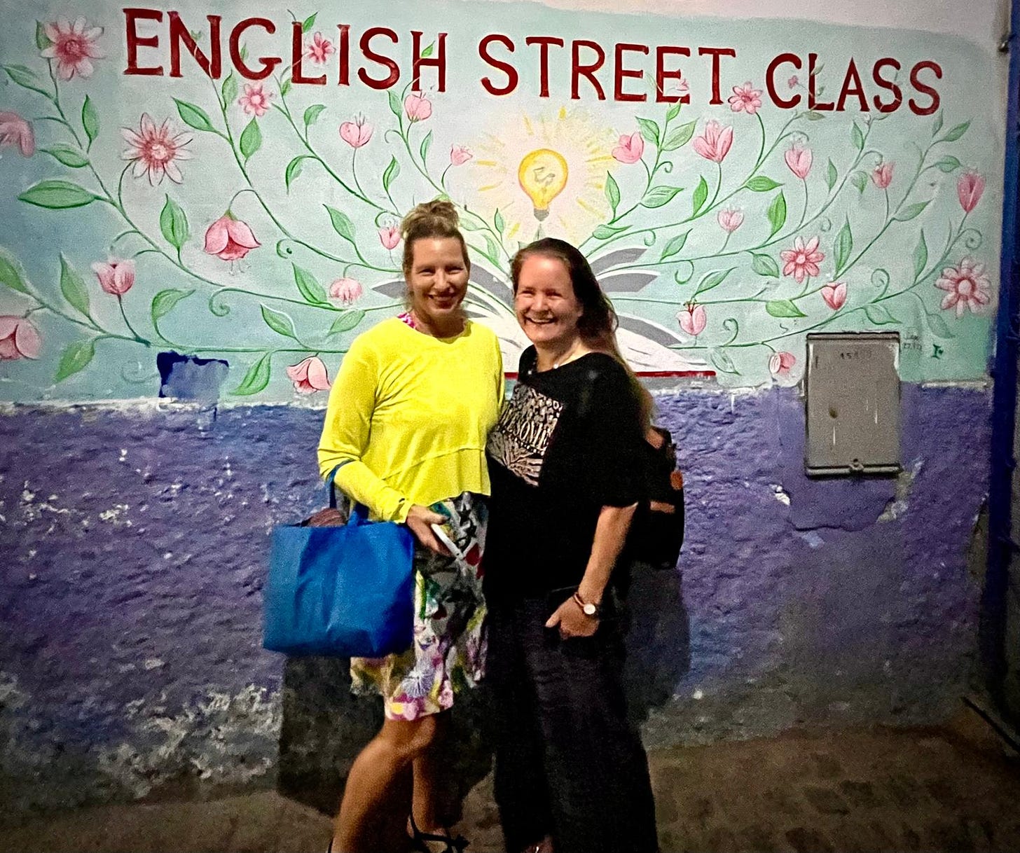 Two white women, one with her hair up wearing a yellow top and one with her hair down, wearing a t-shirt and hugging and smiling at the camera in front of a brightly painted wall. Two white women, one with her hair up wearing a yellow top and one with her hair down, wearing a t-shirt and hugging and smiling at the camera in front of a brightly painted wall.