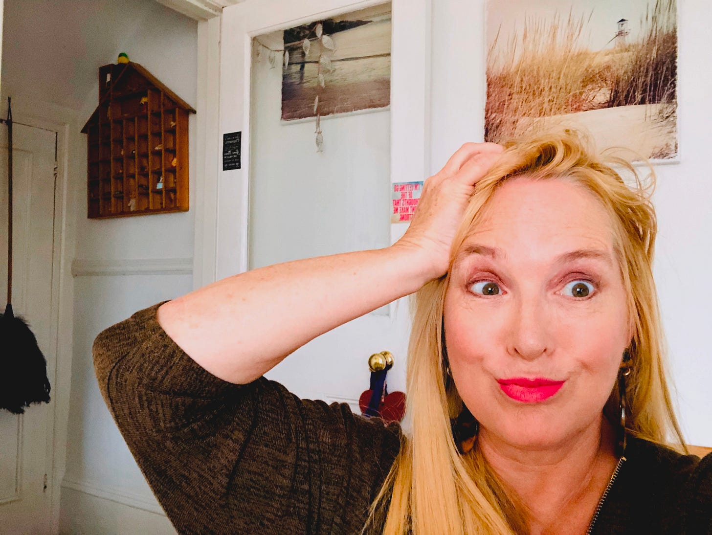 Danusia Malina-Derben pulling a comic face with one hand on her head, inside a home with beachy wall art and a wooden shelf in the background—capturing the chaos-meets-composure energy of creative life.