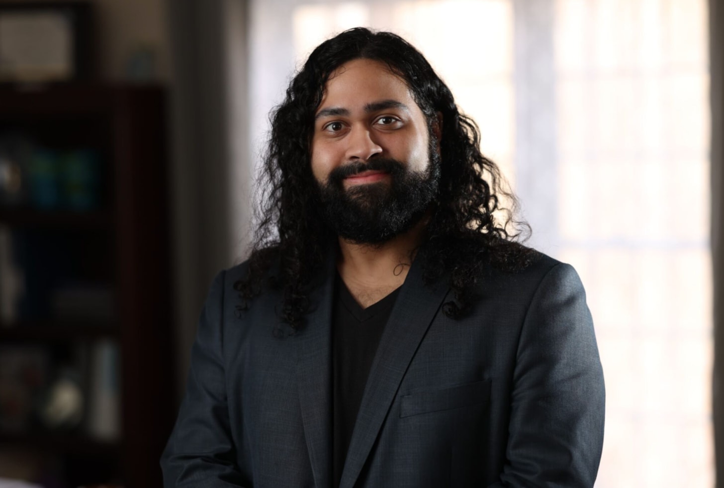 An indigenous man with black, shoulder length hair and a black beard smiles in a gray suit jacket and black shirt.