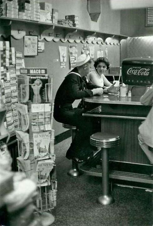 This may contain: a man and woman sitting at a counter in a store with soda cans on the shelves