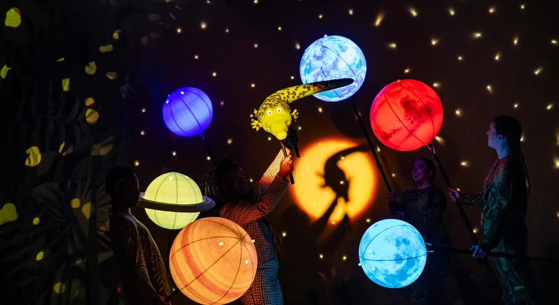 A crocodile puppet with colourful spheres around it and a darkly lit stage