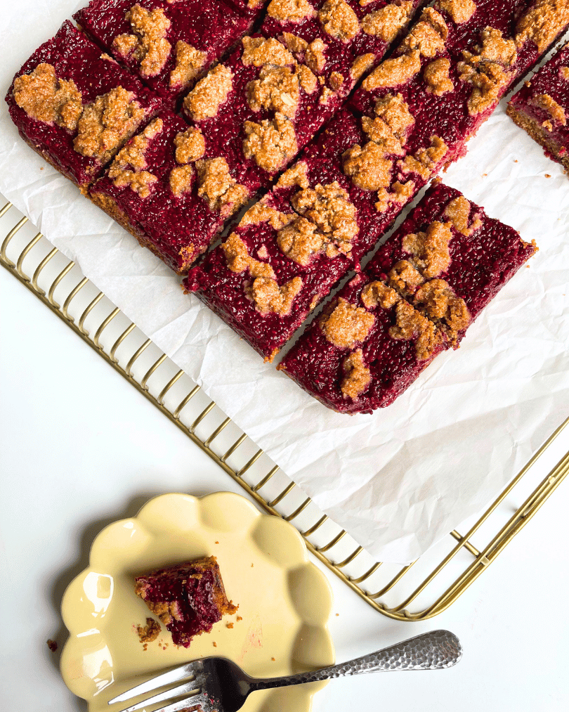 raspberry jam oat bars on a white parchment paper on a gold bake rack, a small yellow plate with a half eaten bar on it.