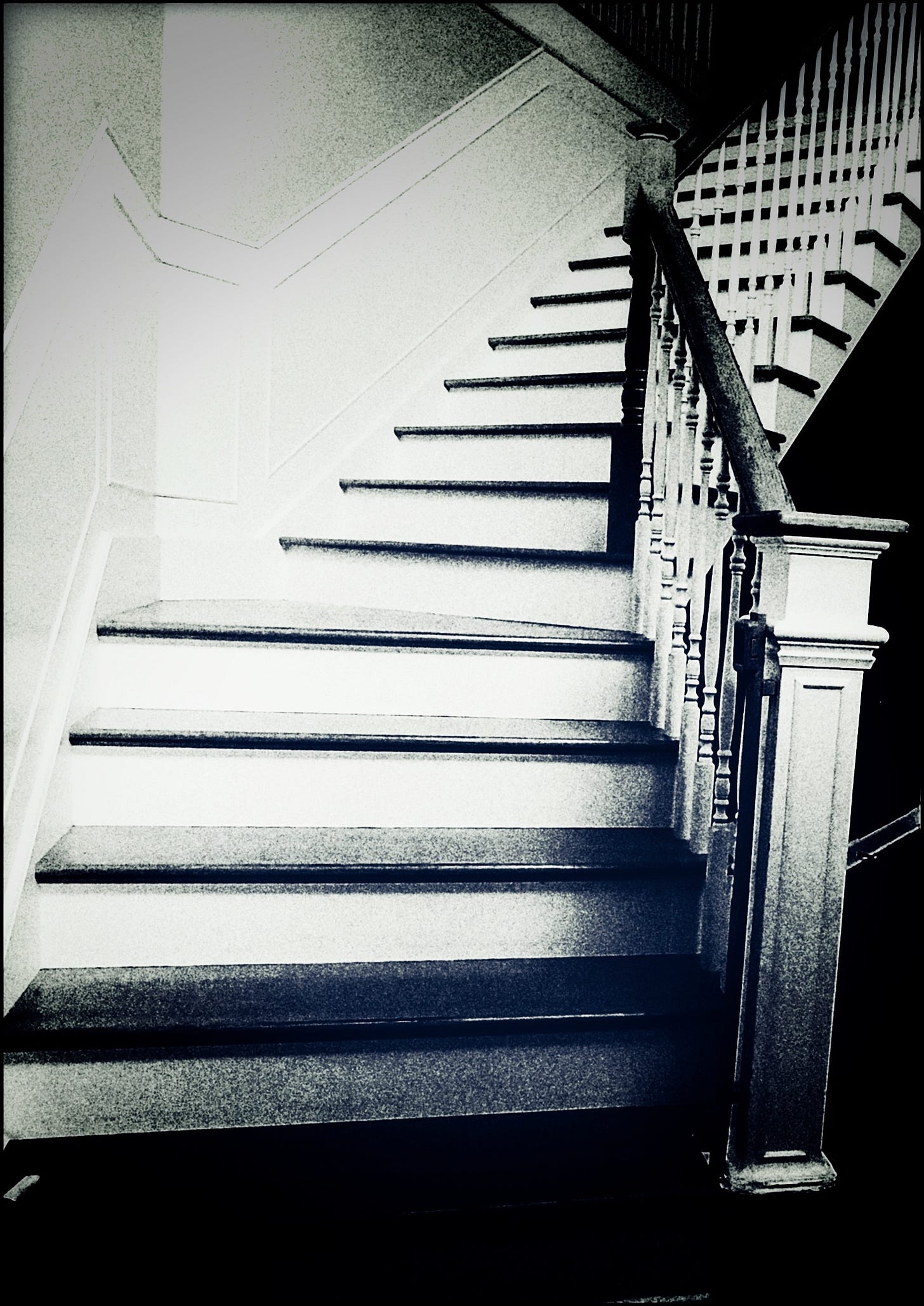 A dim, grainy photo of a banistered wooden stairway in what appears to be an entry foyer. The steps are white; the actual treads are unpainted. The walls are bare. The suggestion is that this is a vacant house.