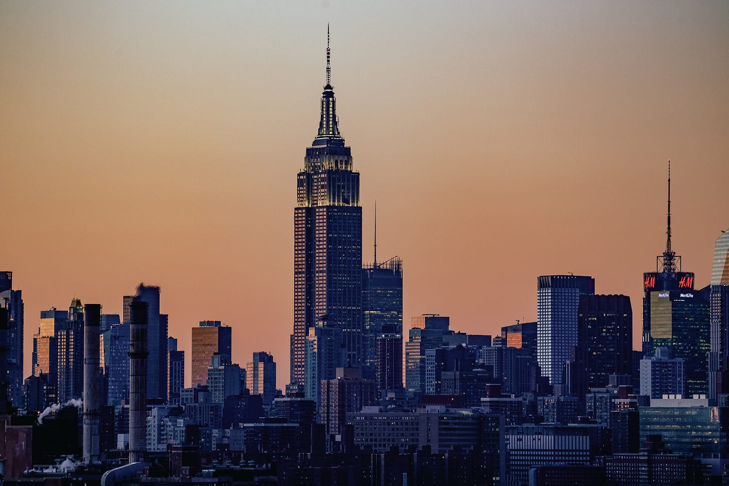 A detailed, medium shot of the Empire State Building standing prominently among the Midtown Manhattan skyline at dusk. The sky is a soft gradient of orange and grey. Many of the surrounding buildings are lit up, and smoke or steam can be seen rising from some of the rooftops in the foreground.