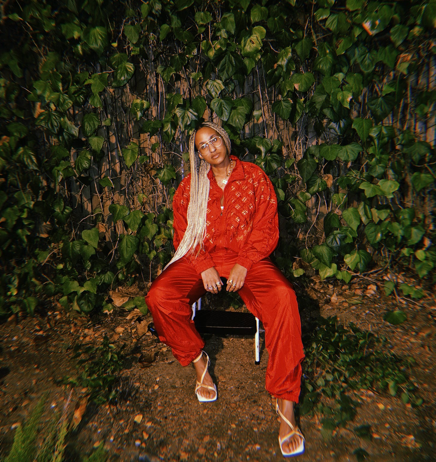 Dr. Cadet sits in front of lush green ivy wearing a red tracksuit from her new vintage collection with yellow heels. With gold details on the tracksuit her golden braids fall to one side she looks stunningly sad yet stylish. 