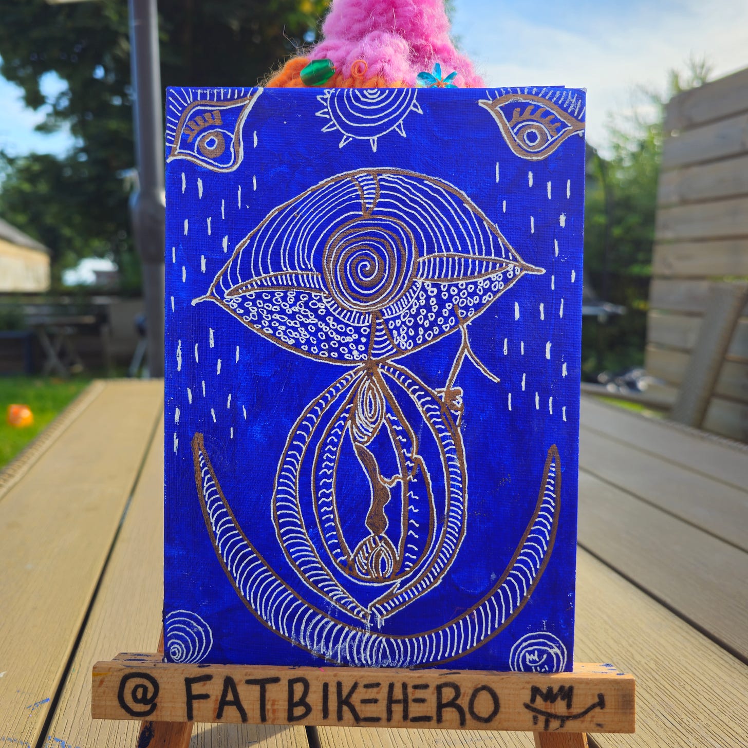 Alt Text: A vertical painting on a blue canvas by Fatbikehero. The artwork features complex gold and white line drawings depicting a large stylized eye with a spiral pupil above a stylized vulva shape. Rain-like dashes fall from the top of the canvas. The painting rests on a small wooden easel signed "@FATBIKEHERO". Alt Text: A vertical painting on a blue canvas by Fatbikehero. The artwork features complex gold and white line drawings depicting a large stylized eye with a spiral pupil above a stylized vulva shape. Rain-like dashes fall from the top of the canvas. The painting rests on a small wooden easel signed "@FATBIKEHERO".