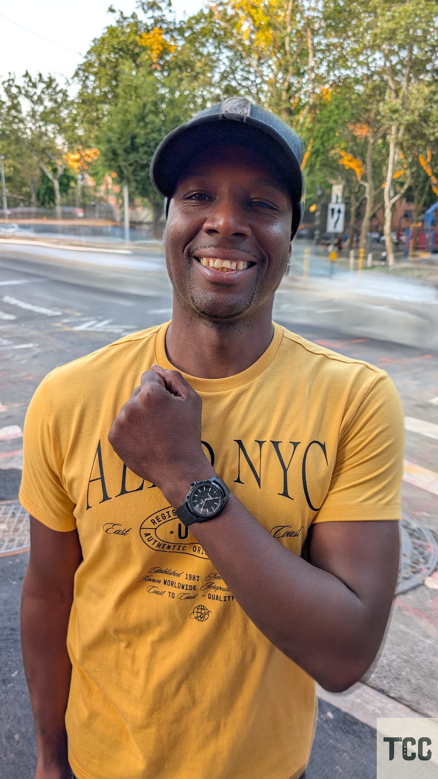 A smiling man standing outdoors wearing a yellow "AERO NYC" shirt and black cap, proudly raising his wrist to show a Formex Reef watch. Trees and street blur are visible in the background, captured with a long exposure effect.