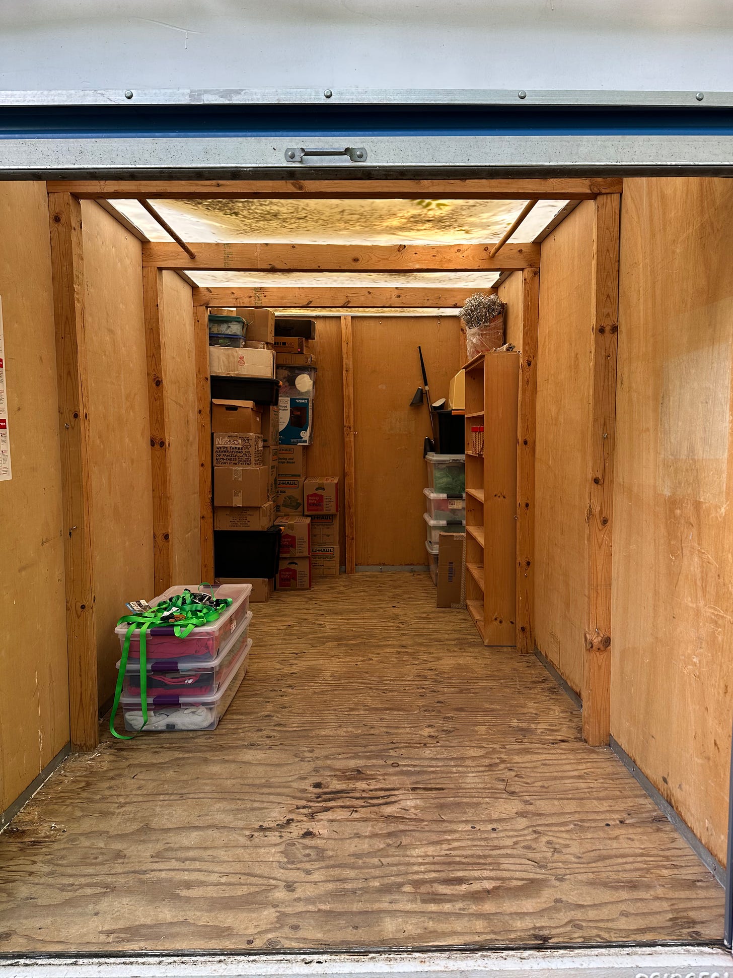 A view inside a trailer with wooden sides and floor, with a high stack of boxes, a lamp, and a bookcase visible in the background, with a stack of plastic bins and green straps hanging off them.