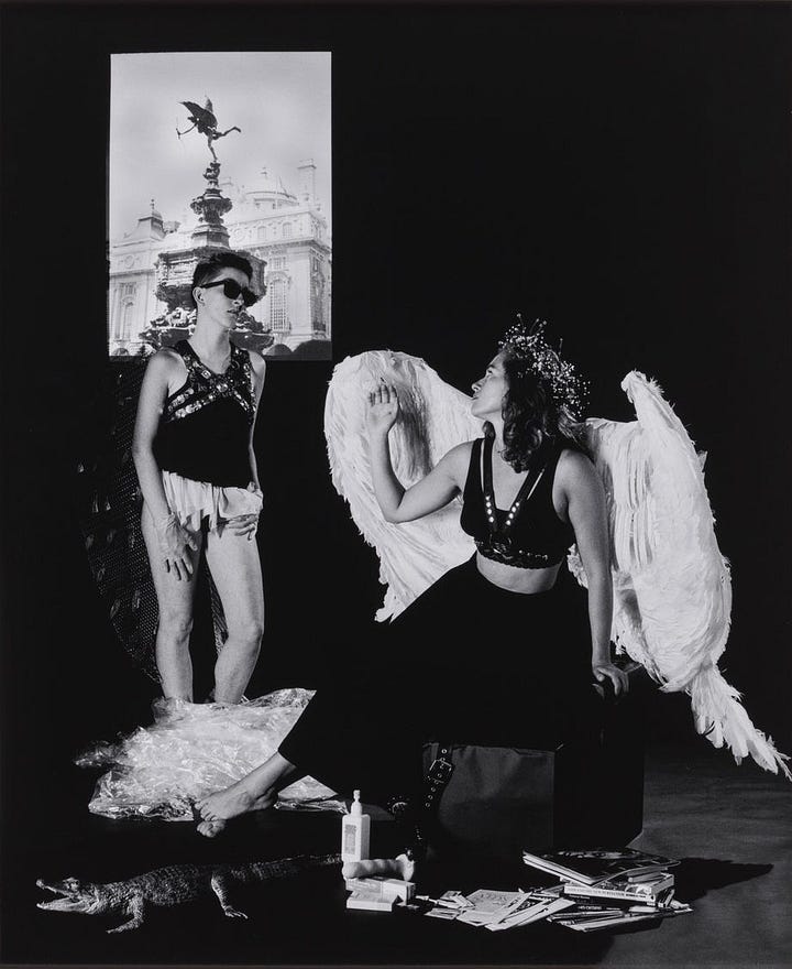 1) A black-and-white photo of a barefoot person in large white angel wings and a floral crown, sitting on a box and reading a magazine. On the wall behind them, there is a black and white photograph of angelic statues. In front of the portrait, there is another angel wrapped up in clingwrap. There is a faux alligator on the floor, surrounded by safe sex magazines and pamphlets. There is also a dildo and a bottle of lotion on the floor, in front of the angel. The scene feels surreal. 2) Two people in elaborate costumes face each other. One is sitting wearing large, white angel wings, while the other stands. A statue photo is visible behind the person who just emerged from the clingwrap. The clingwrap sits around their feet on the ground, next to the faux alligator. The person who was wrapped up is wearing a leather chest harness, a long frilly shirt covering their crotch, sunglasses, and latex gloves on both hands. They also have dark angel wings, with pieces of metal hanging off of them in several places. The person sitting down is looking over their wings to look at the other angel. The floor in front of them is still full of magazines, pamphlets, books, and a dildo with some lotion/lube. This photo is in black and white.