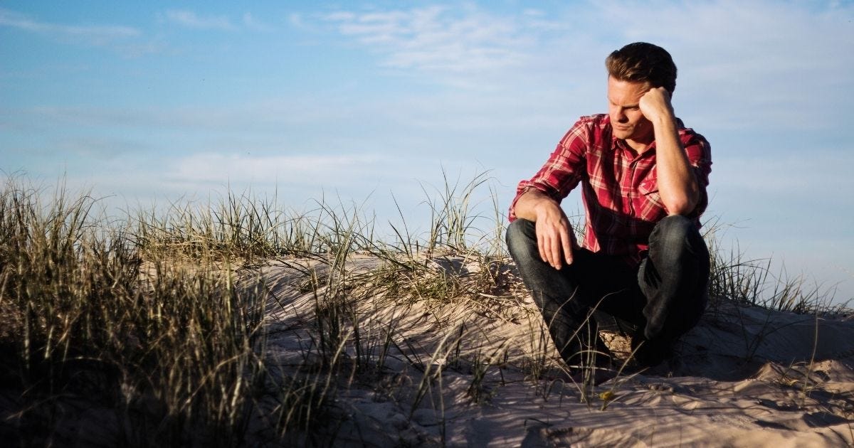 Man in a red plaid shirt sitting thoughtfully on a sandy dune in Australia, surrounded by grass