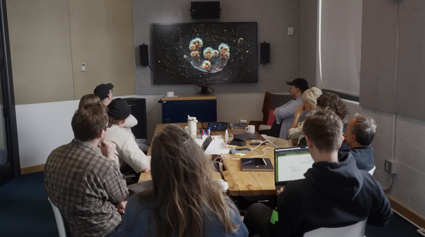 A group of people sitting in an office around a table as they look at a small monitor displaying generative AI footage of tadpoles.