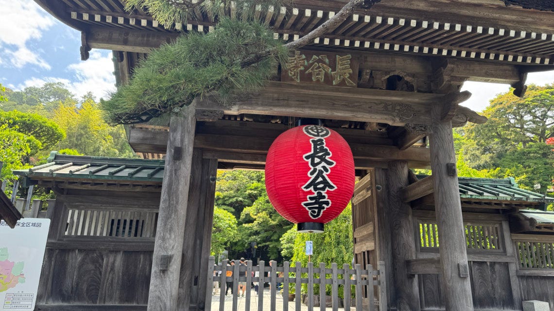 Wooden temple gate with a red lantern reading “長谷寺” (Hase-dera) surrounded by lush greenery and trees.