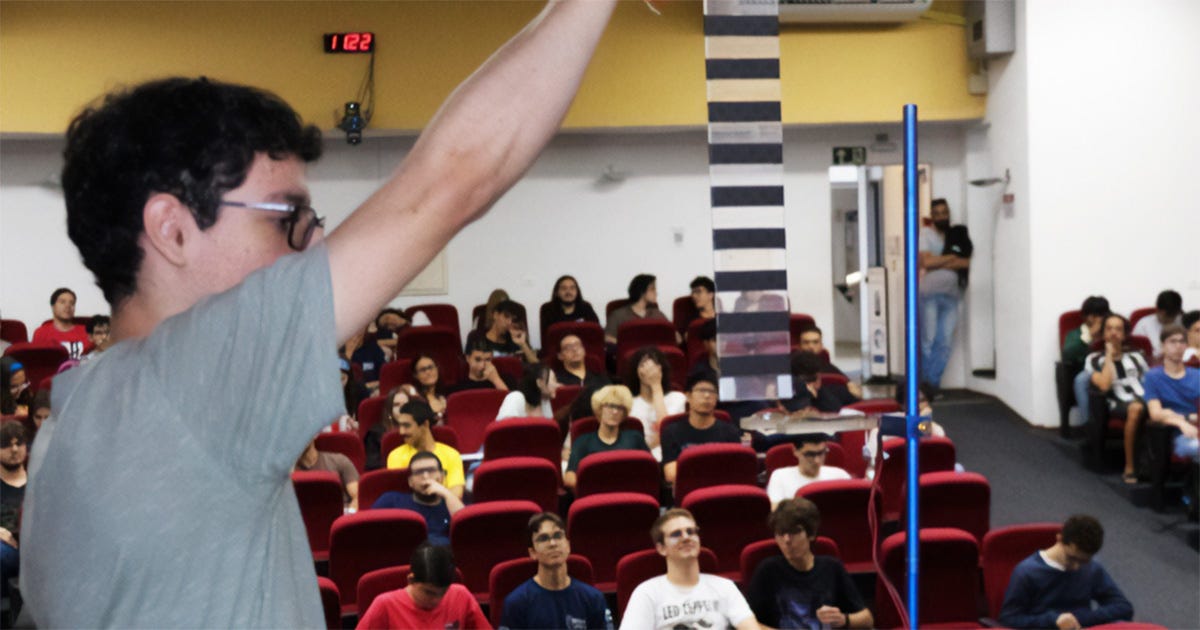 Fotografia de uma demonstração experimental em um auditório. Em primeiro plano, uma pessoa segura uma régua listrada acima de um aparato de queda livre com sensor óptico. Ao fundo, dezenas de estudantes sentados acompanham a atividade, observando o experimento.