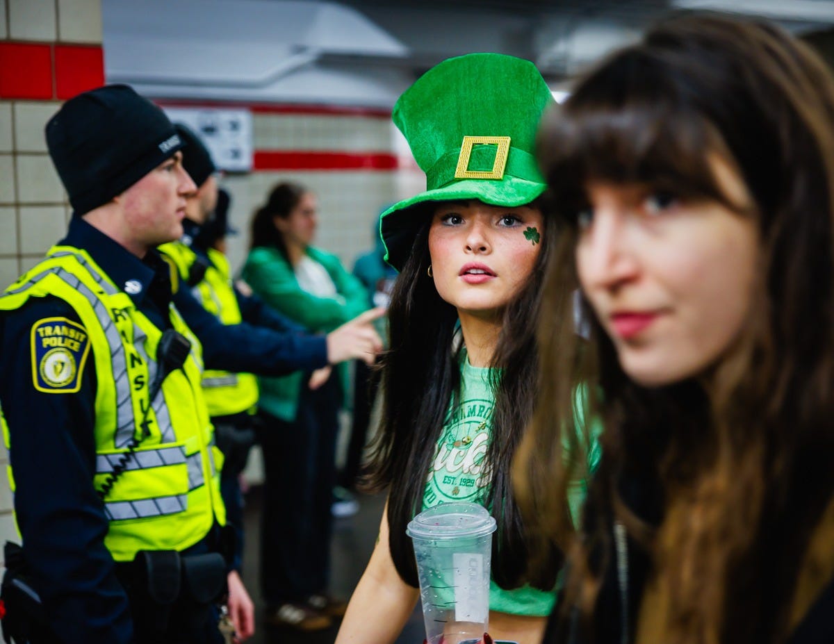Young woman wearing a large green St. Patrick’s Day hat looking toward the camera inside a Boston subway station, with police officers in the background. Young woman wearing a large green St. Patrick’s Day hat looking toward the camera inside a Boston subway station, with police officers in the background.