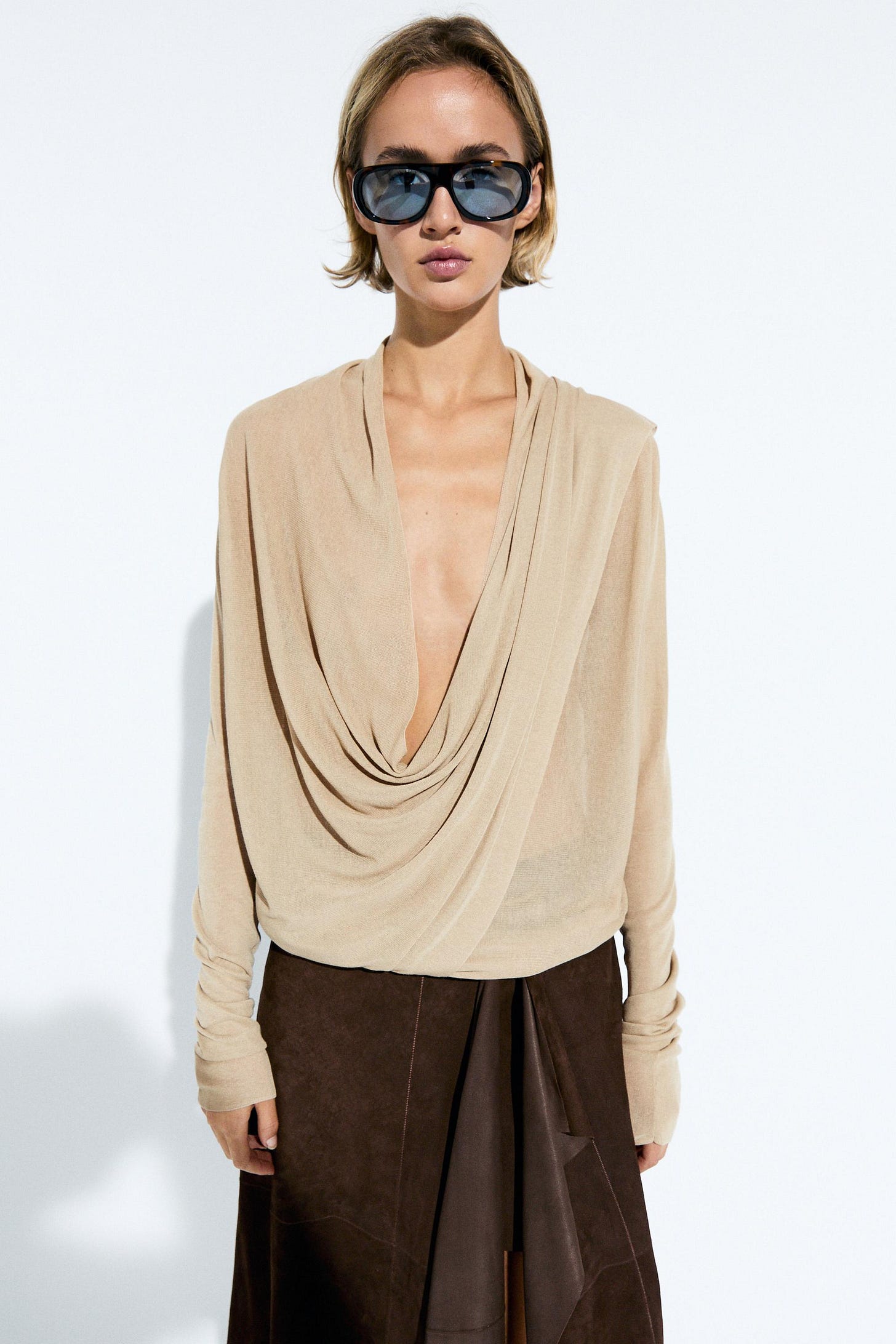 Ensemble composé d'un gilet couleur sable à décolleté drapé profond et manches extra-longues, d'une jupe marron et de lunettes de soleil.