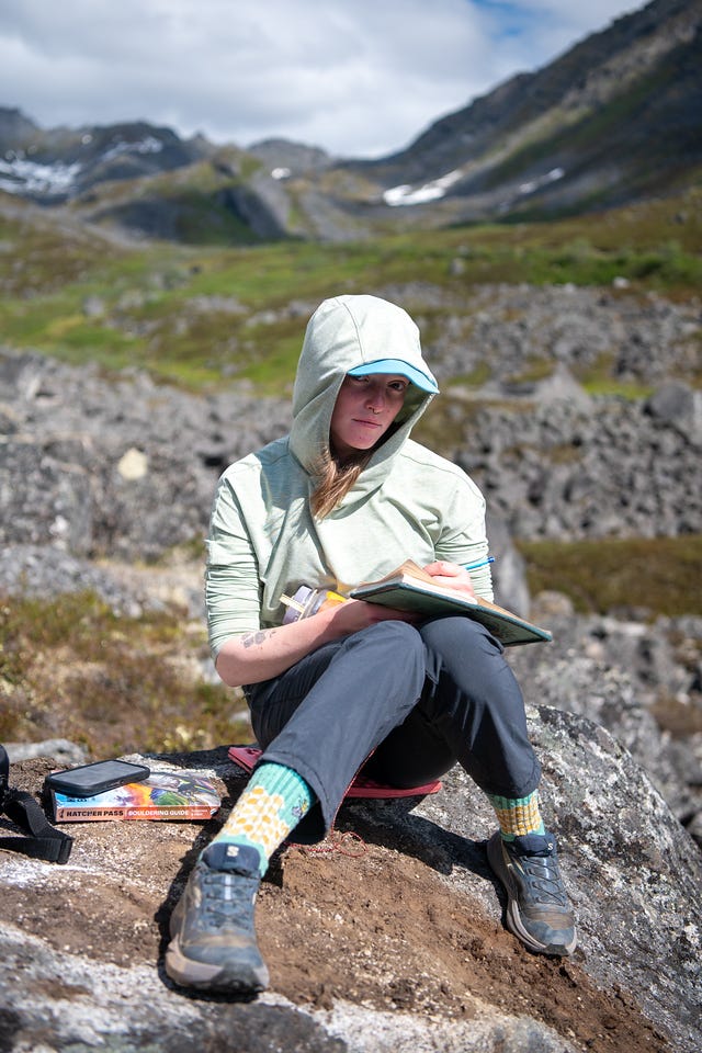 A woman sits on a rock with a small notebook open on her knee, pen in hand, looking up at the camera from under the hood of a pale green sun hoody. Yellow-patterned socks show above trail shoes. Beside her on the rock sit a phone and a Hatcher Pass bouldering guidebook. Behind her, a boulder-strewn green valley rises to dark ridgelines holding streaks of snow. A woman sits on a rock with a small notebook open on her knee, pen in hand, looking up at the camera from under the hood of a pale green sun hoody. Yellow-patterned socks show above trail shoes. Beside her on the rock sit a phone and a Hatcher Pass bouldering guidebook. Behind her, a boulder-strewn green valley rises to dark ridgelines holding streaks of snow.