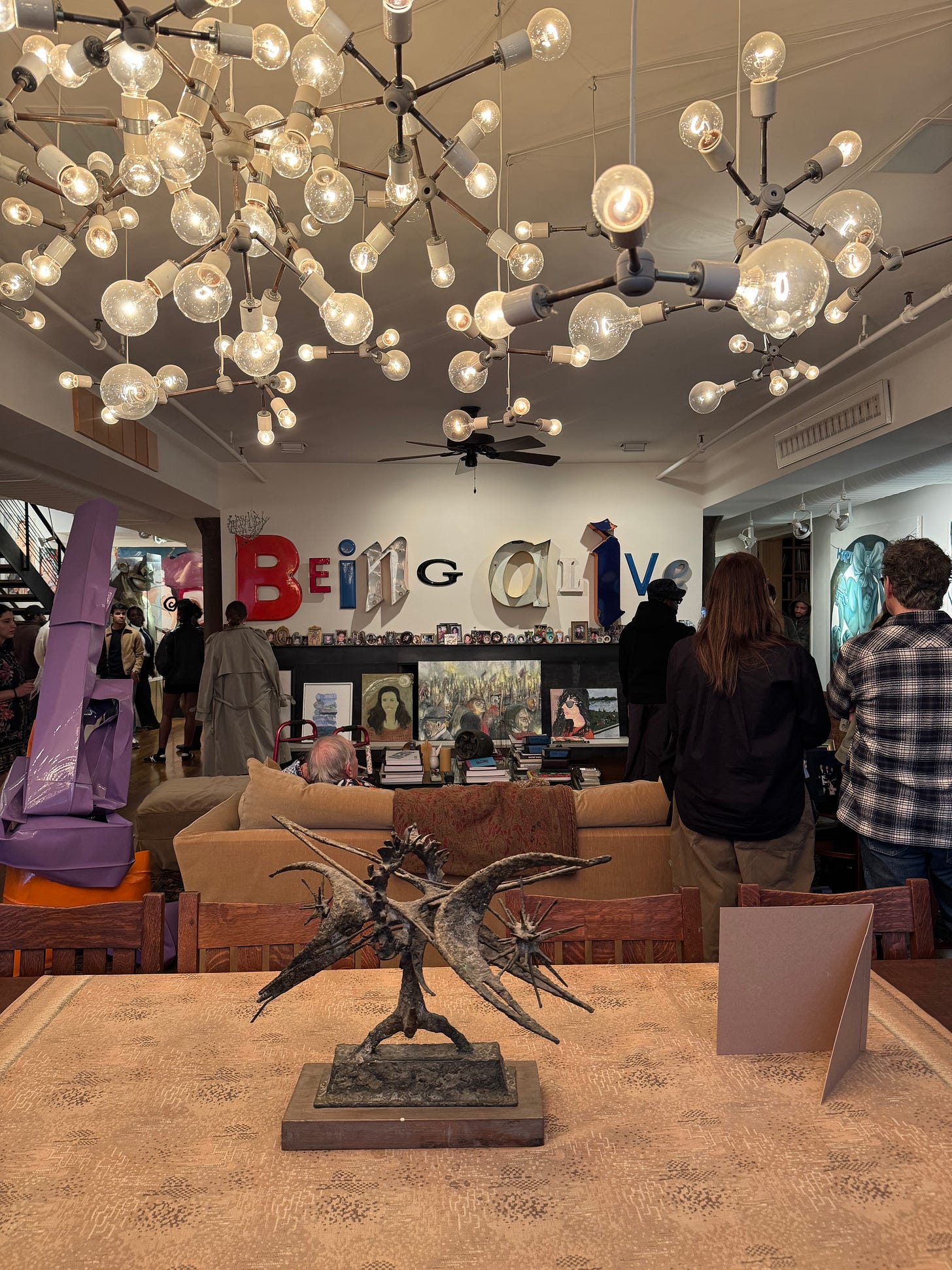 The living room of the Hort Family Collection during New York Art Week, filled with visitors, vintage furniture, sculptures, artworks, and a branching chandelier made from exposed bulbs.