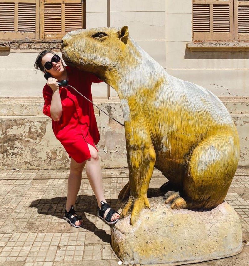 Fotografia de Manu Barem, de vestido vermelho e em pose dramática, atendendo ao telefone num orelhão em formato de capivara gigante no meio da rua.