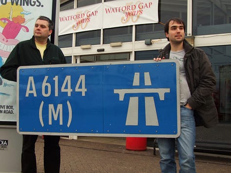 Three photographs showing people posing with the A6144(M) sign in various settings - on the left in front of a building; in the middle, two people obscured by the signs and clearly having trouble holding them; on the right a whole group posing together with the sign in a lay-by