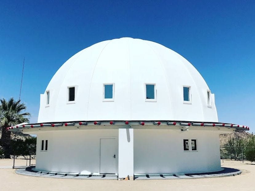 Image: A white domed structure in the desert surrounded by sand a blue sky.