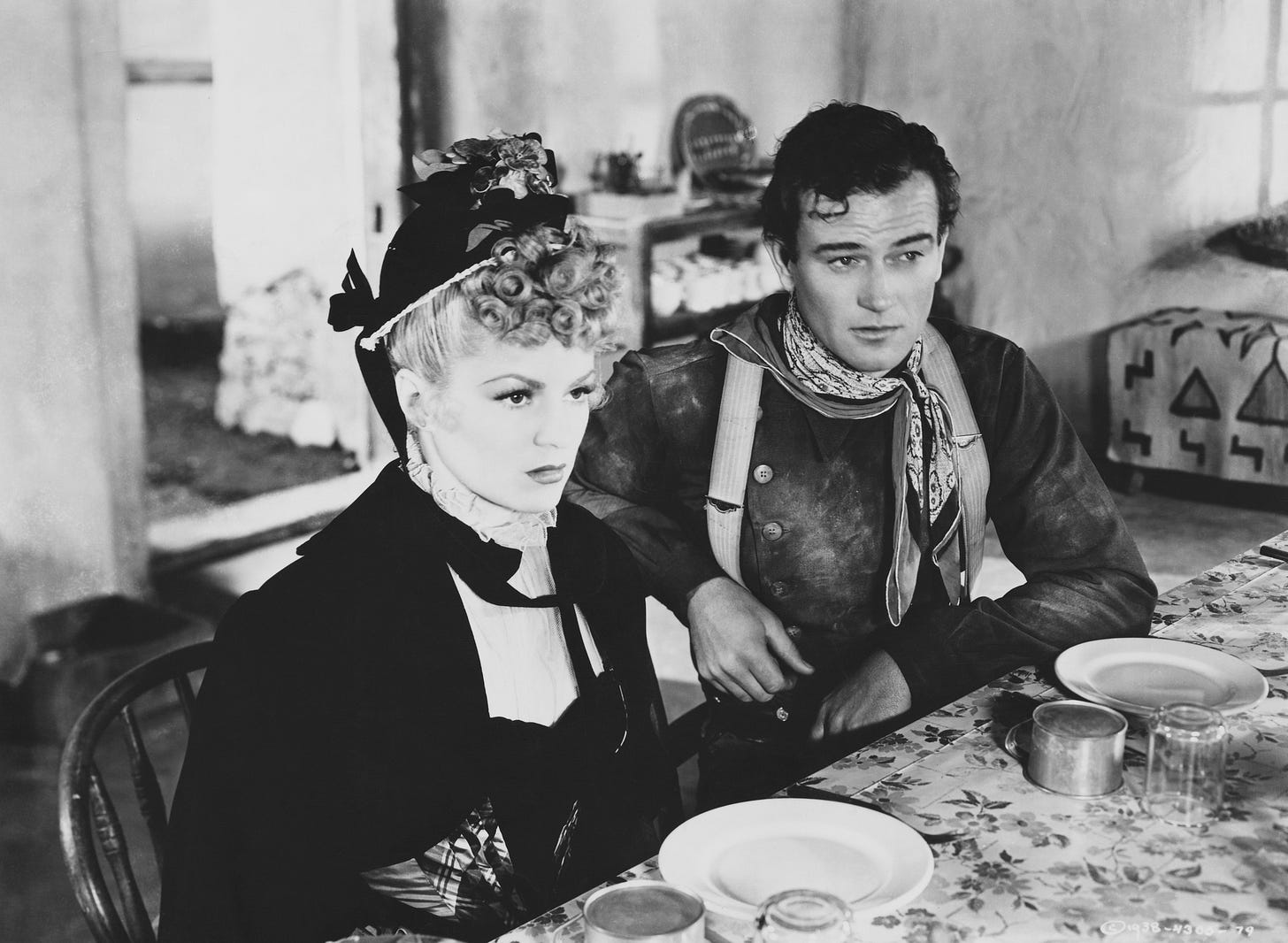 Claire Trevor in a black hat and dress and John Wayne in what looks like a Union frock coat sit at a dining table in "Stagecoach" (1939)