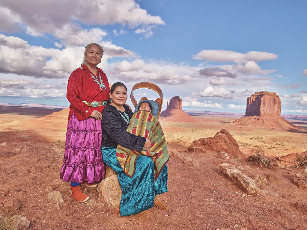 Three generations of Navajo women, a grandmother standing and a mother seated holding an infant child – all in traditional dress outdoors in Monument Valley Navajo Tribal Park.