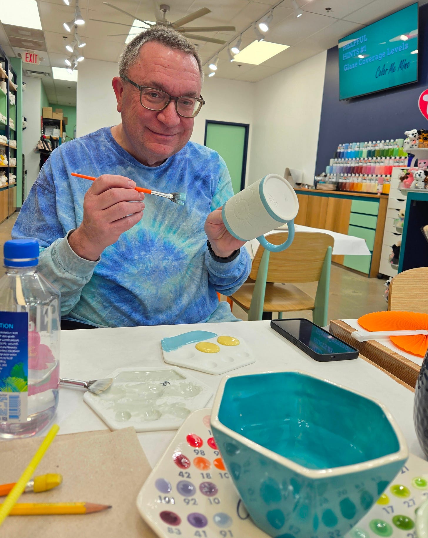 John painting a mug at Color Me mine. paint brush in one hand and mug in the other. He is smiling for the camera.