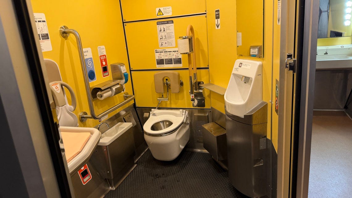 A spacious, yellow-walled accessible train restroom equipped with a toilet with grab bars, a urinal, a lower sink, and a wall-mounted baby seat. A spacious, yellow-walled accessible train restroom equipped with a toilet with grab bars, a urinal, a lower sink, and a wall-mounted baby seat.