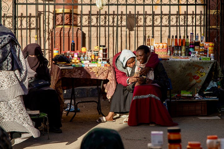 Top left: A busy market street in Aswan with vendors and shoppers beneath a mosque minaret and tangled overhead wires Top right: Two women sitting together sharing a phone beside a table of cosmetics and bottled goods, with a woman in a niqab seated nearby Bottom right: An elevated view across the rooftops of old Aswan toward the Nile, with feluccas and cruise ships moored along the banks and the modern city beyond Bottom left: A young man crouched over an open telecommunications junction box with tangled colored wires, while a woman in a pink hijab and a shopkeeper stand in the doorway behind him