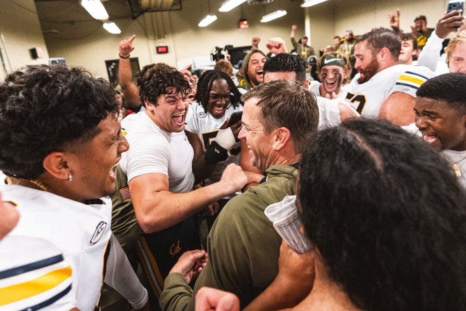 First image shows a group of male athletes in white and blue uniforms huddled excitedly in a brightly lit indoor locker room area with overhead lights and some equipment visible, several players smiling and embracing a central figure in green attire. Second image depicts two male players in blue and gold uniforms standing close on a green football field with a large crowd of spectators in red and white in the stadium stands behind them, one player with number 81 on jersey gesturing animatedly. Third image captures multiple male athletes in blue and gold uniforms on the field at night under stadium lights with a crowd in background, players in helmets and pads celebrating with arms raised near the sideline. Fourth image features a cluster of male athletes in white and blue uniforms gathered closely in an indoor setting with some wearing caps and holding cameras, expressing joy through smiles and gestures amid overhead lighting.