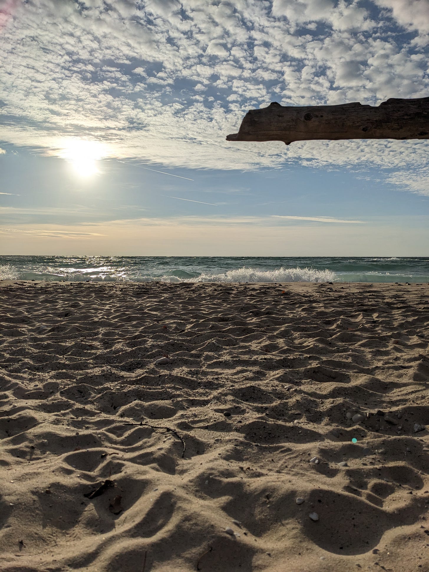 The shoreline of Lake Michigan. There is sand in the foreground, with waves running up to the beach. The sky is blue with scattered white clouds, and the sun is hanging directly ahead at the horizon line, shining in a fuzzy burst of white light.