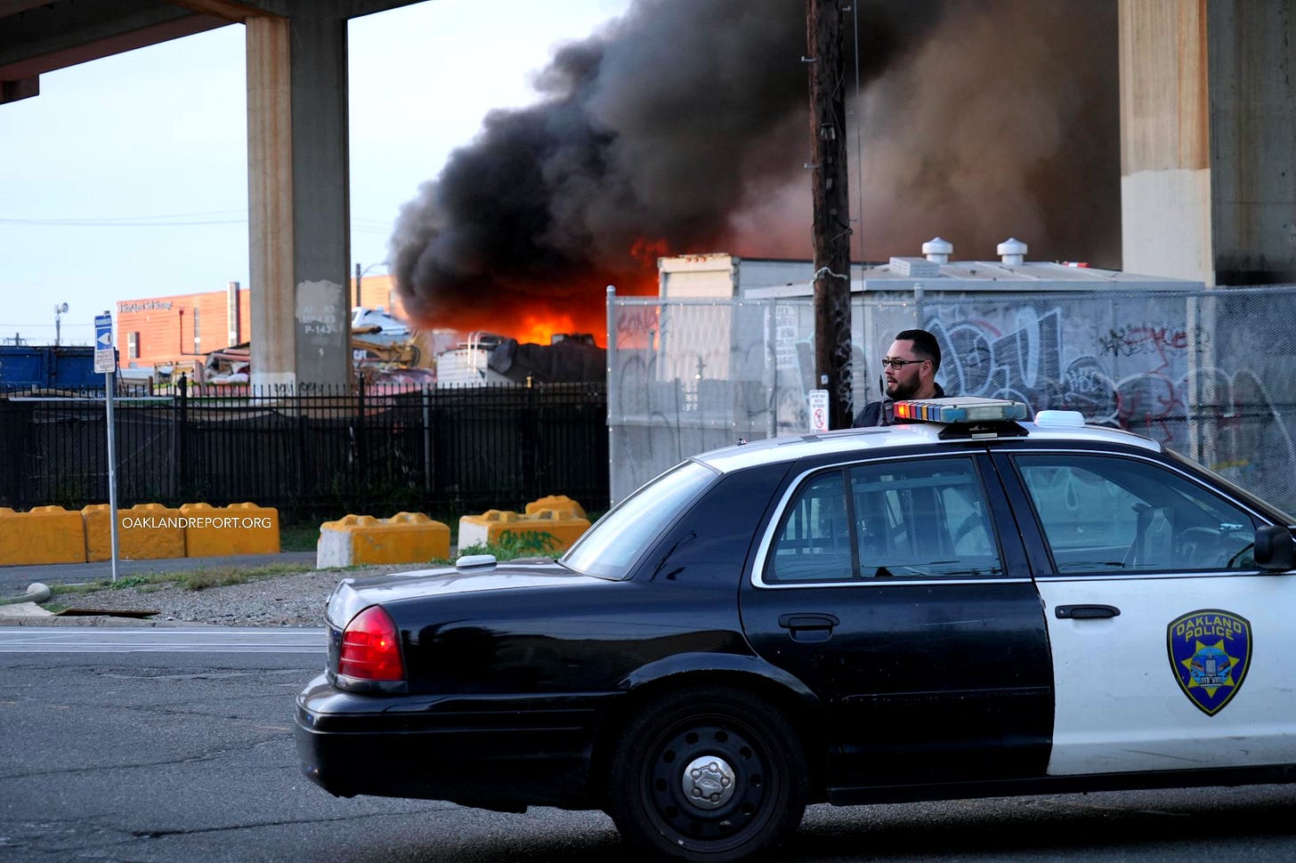 A police officer outside at a transfer station fire in East Oakland on Apr. 28, 2026. Two RVs caught fire in the enclosed lot. (Photo: Shutterstock / Oakland Report) A police officer outside at a transfer station fire in East Oakland on Apr. 28, 2026. Two RVs caught fire in the enclosed lot. (Photo: Shutterstock / Oakland Report)