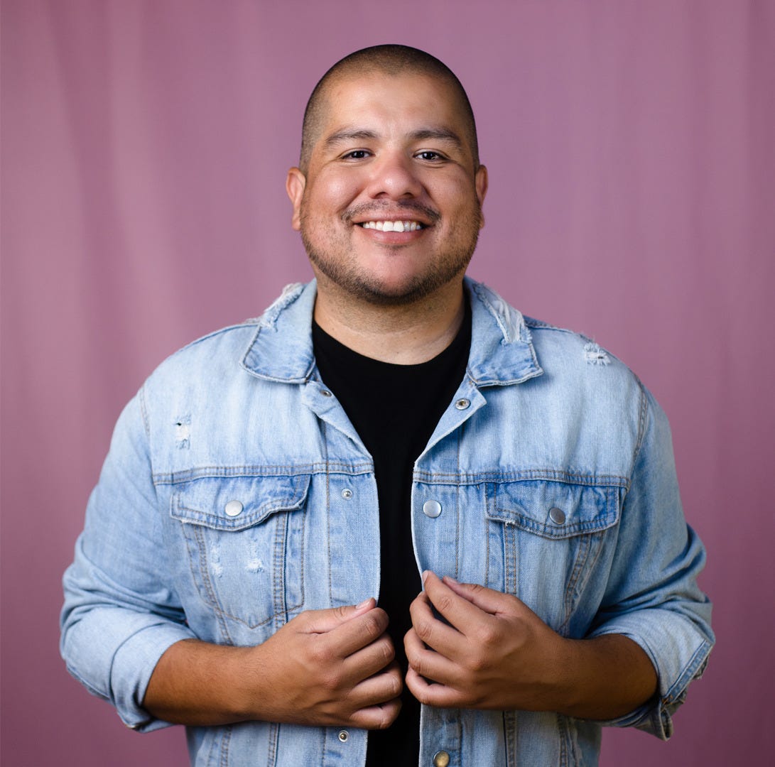 In this photo, Julio is a smiling Hispanic man standing against a light pink background, wearing a light blue denim jacket over a black t-shirt. His hands grasp the jacket sides, conveying a friendly and approachable demeanor. He is smiling straight into the camera, he has mustache and beard stubble, and a shaved head.