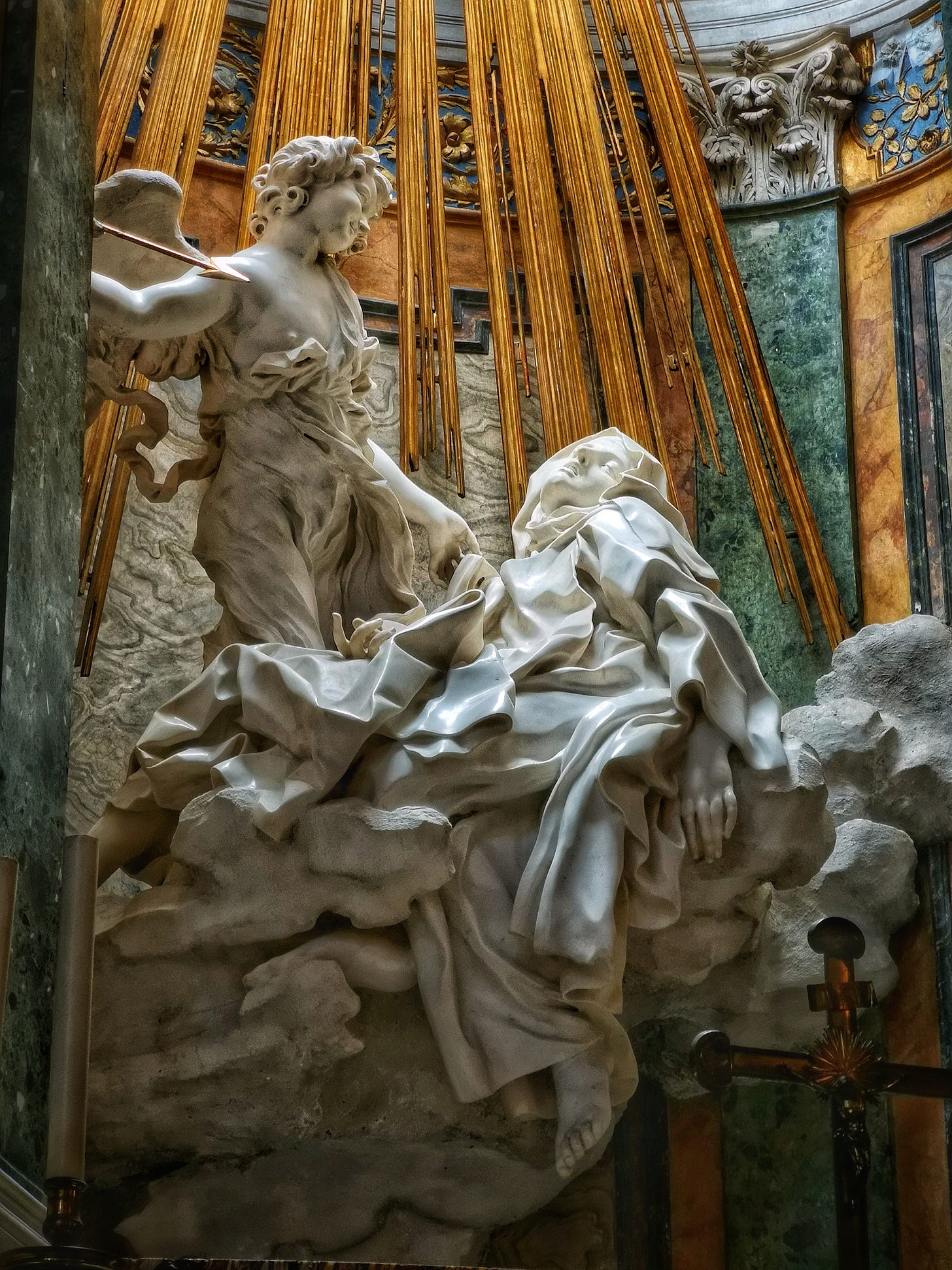 A detailed photograph of Gian Lorenzo Bernini's Baroque marble sculpture, "The Ecstasy of Saint Teresa." An angel holds a golden arrow, smiling as he looks down at Teresa. She is swooning on a cloud base, head thrown back, eyes closed, and mouth slightly open in an expression of overwhelming spiritual and physical rapture. Golden rays of divine light descend behind them against colored marble walls.