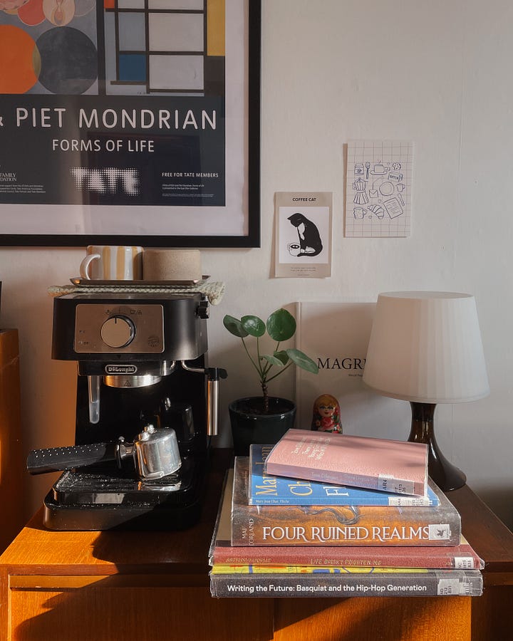 March TBR - Image 1: A stack of library books on my coffee station, with a coffee machine and decor in the background. / Image 2: A close-up of the book stack.