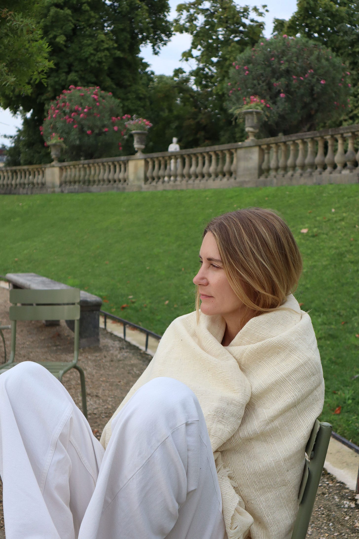 Woman with shoulder-length blonde hair in profile, wrapped in a cream woven blanket, looking out over a garden with pink flowers and a stone balustrade. Woman with shoulder-length blonde hair in profile, wrapped in a cream woven blanket, looking out over a garden with pink flowers and a stone balustrade.