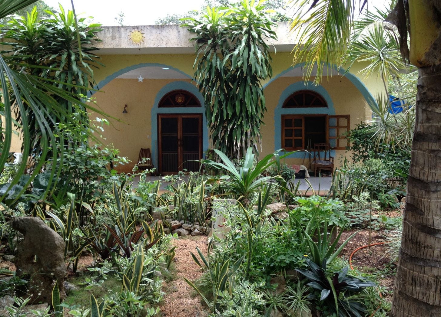 Yellow cinder block house with blue-trimmed arches, a painted sun over the door, a white star hanging from the porch, garden in front. Yellow cinder block house with blue-trimmed arches, a painted sun over the door, a white star hanging from the porch, garden in front.