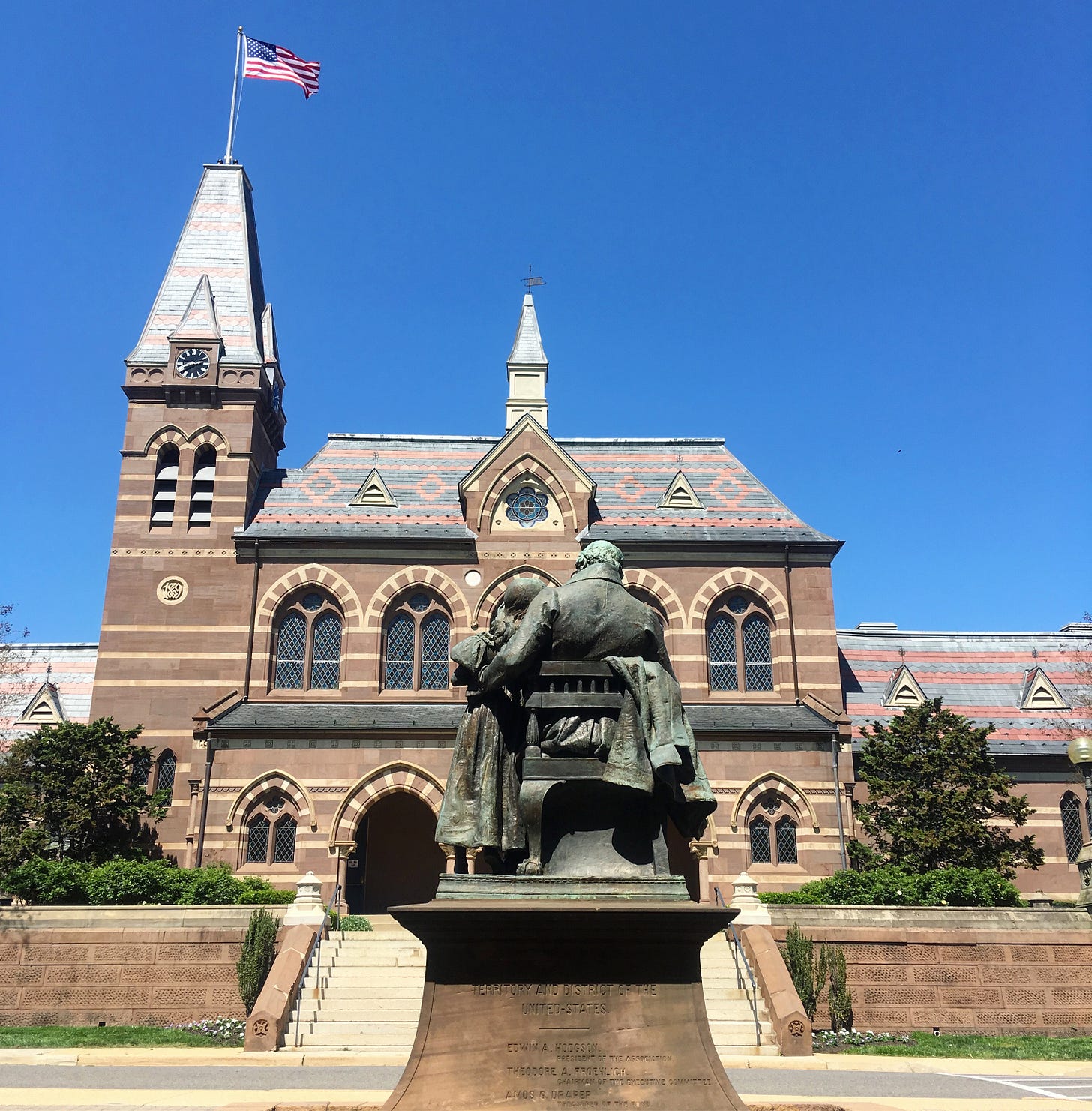 A shot of the Thomas Gallaudet statue at Gallaudet University. A brick striped university hall stands in the background, up a flight of stairs, and with a tower and American flag on the left-hand side of the image. Bright blue skies in the background.