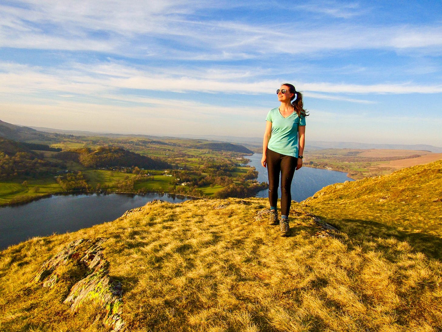 A woman hiking stands on a fell summit in the Lake District, with a lake and green valley stretching out behind her under a blue sky. A woman hiking stands on a fell summit in the Lake District, with a lake and green valley stretching out behind her under a blue sky.
