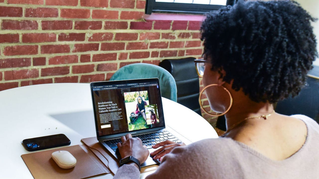 Black woman working on laptop, viewing website, seated at table with phone and mouse, brick wall background