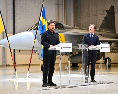 Zelenskyy and Swedish PM Ulf Kristersson speak at podiums in front of military jet in hangar