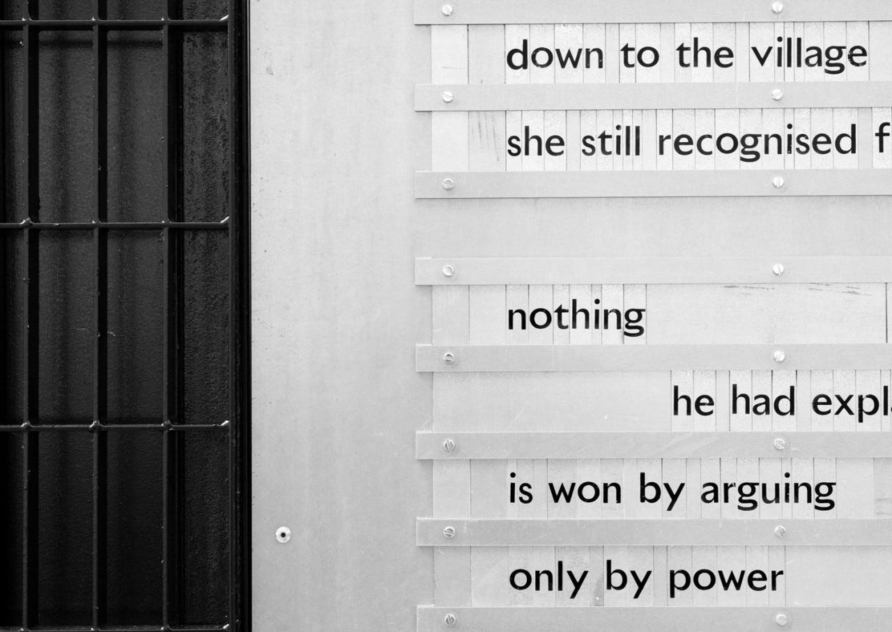 Words set as individual letters in rows on a wall by a window covered with a grille
