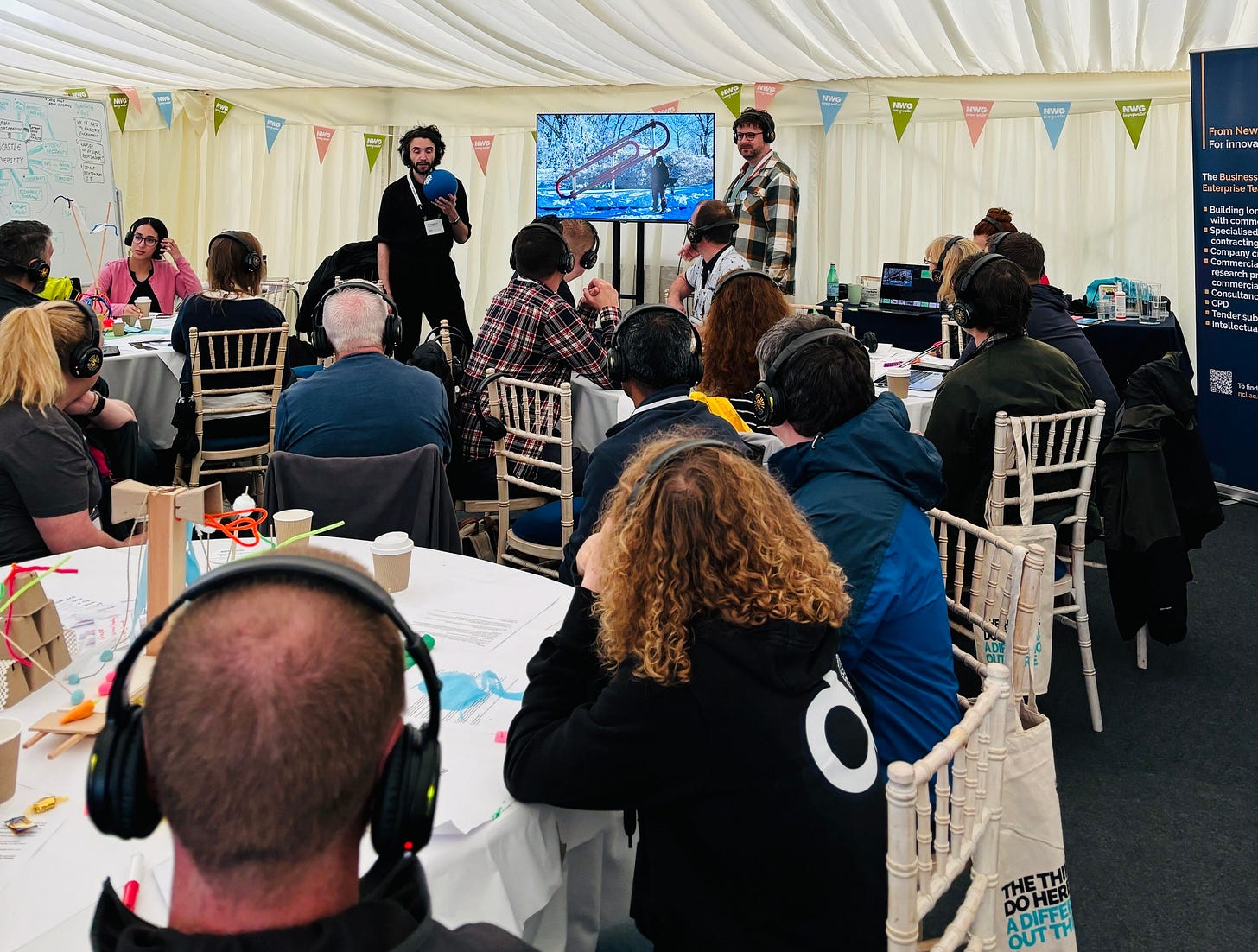 A marquee tent filled with people sitting in white painted, faux bamboo chairs around large white table clothed tables. Attendees are watching two men presenting at a screen. The screen shows a photo of the world's largest paperclip. The attendees are a diverse bunch. The speakers (Alex and Andrew) are not.
