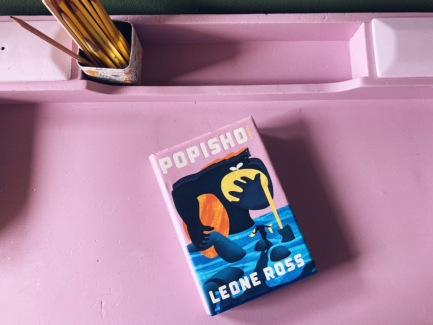A novel called Popisho by Leone Ross sits atop a desk painted a shade of purple to match the novel's cover