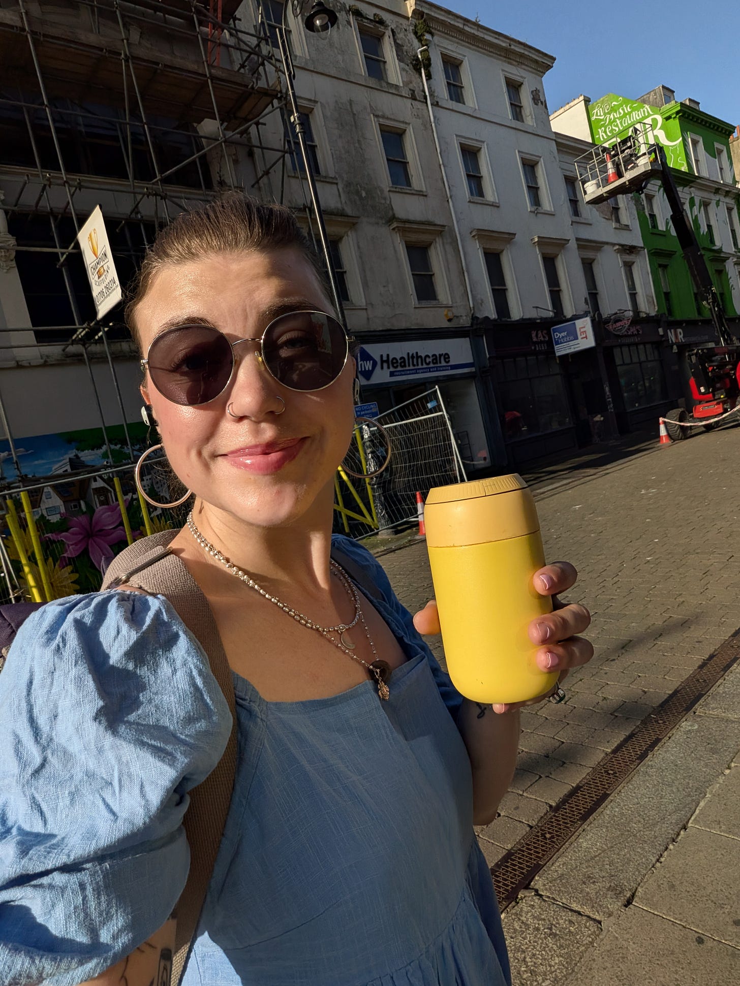 Grace smiling goofily while holding her yellow coffee flask; prescription sunnies on, lip gloss in check, hair scraped back and blue summer dress on as well as her backpack. There's scaffolding on the rotting buildings behind her as she's walking through the worst part of town, but the sun is glowing on her face so all is well.