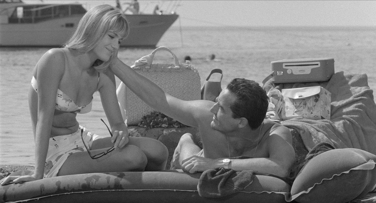 la foto in bianco e nero mostra Catherine Spaak e Vittorio Gassman in un celebre scena de film: sono seduti su un materassino su una spiaggia, lui sdraiato prono le accarezza i viso, lei seduta lo guarda e sorride